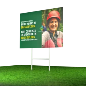 A billboard on grass shows a smiling person in a red helmet and outdoor gear. The sign reads: “Adventure is waiting. Build yours at beascout.org. Aquí comienza la aventura en beascout.org.”.