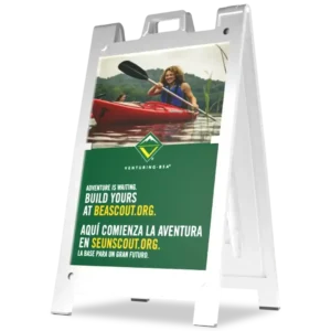 A sidewalk sign displays an image of a smiling person kayaking on a lake, promoting Scouting adventures with the text: Adventure is waiting. Build yours at beascout.org and its Spanish translation below.