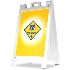 A white sandwich board sign displays the Cub Scouts logo, featuring a wolf’s head inside a yellow diamond with “CUB SCOUTS” text, on a bright yellow background.