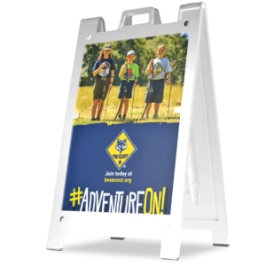 A sidewalk sign features three children in Cub Scout uniforms standing outdoors with hiking sticks. The bottom of the sign displays the Cub Scouts logo, website, and the hashtag #AdventureOn! in bold yellow text.