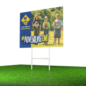 A sign on grass shows three children in scout uniforms holding hiking sticks, with the Cub Scouts logo, website beascout.org, and the slogan #ADVENTUREON! in bold yellow and white letters.