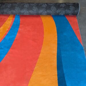 The ShowStopper Event Flooring features bold, wavy stripes in blue, orange, and red. Its top edge is rolled to reveal a gray floral pattern underneath, all displayed on a concrete floor.