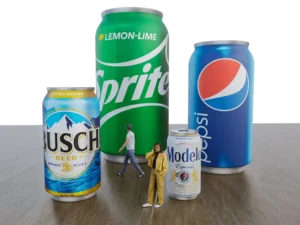 A digitally edited image features three oversized Inflatable Can, Sealed Air products resembling Busch beer, Sprite, and Pepsi cans, with a regular-sized Modelo can. Two miniature people stand and walk among the cans on a wooden surface.