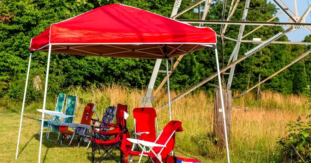 A group of colorful folding chairs sits under a red canopy tent on grass, with tall dry grass, trees, and a metal structure in the background. The scene is outdoors on a sunny day.