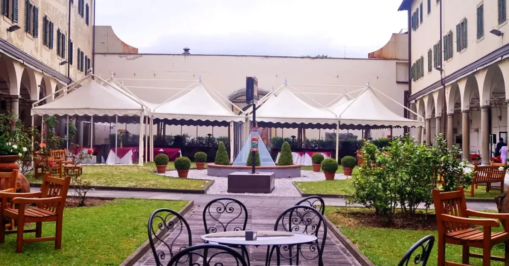 A courtyard with green grass, trimmed bushes, and a central water fountain. White tents and banquet tables line the background; black wrought iron chairs and tables are in the foreground. The scene is bright and inviting.