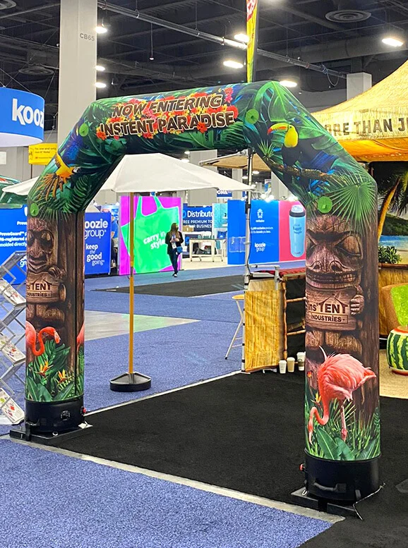 An inflatable arch decorated with tropical designs, tiki faces, and flamingos stands at an event entrance. The arch reads Now Entering Instant Paradise. Booths and banners are visible in the background.
