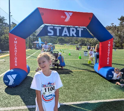 A smiling young girl in a race shirt and bib stands near the starting line arch at a YMCA event on a sunny day. People and children are visible in the background on the sports field.