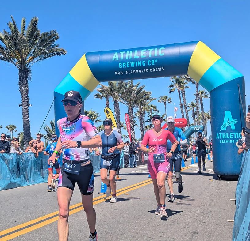Runners in athletic gear approach the finish line under a blue and yellow inflatable arch at a race event. Palm trees and spectators line the street on a sunny day.