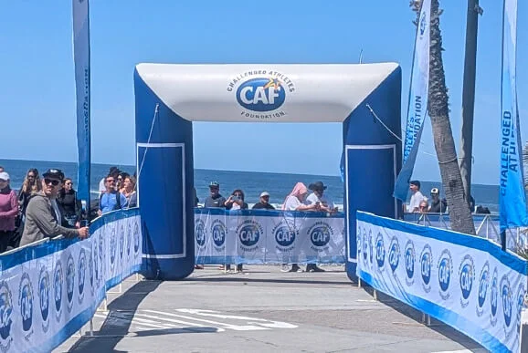 A blue and white inflatable archway marked CAF stands at the finish line of a seaside race, with crowds of people gathered on either side and the ocean visible in the background.