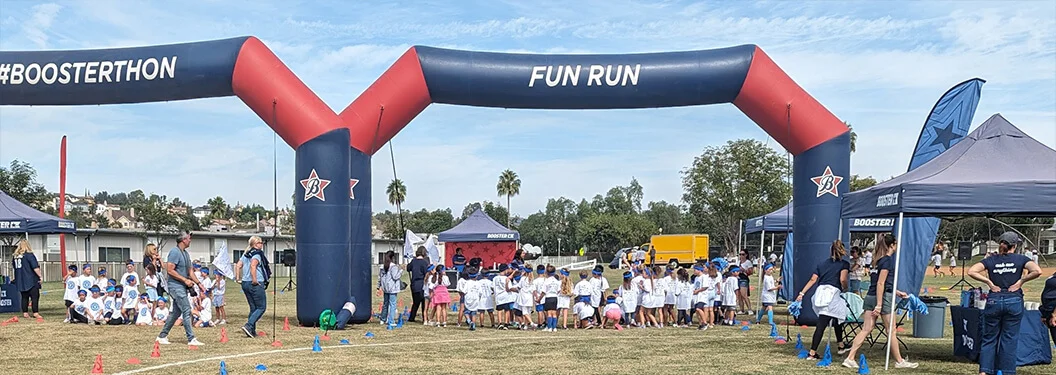 Children in white shirts gather at the starting line under a large inflatable arch reading FUN RUN at an outdoor event. Adults supervise nearby, and event tents are set up on the grassy field.