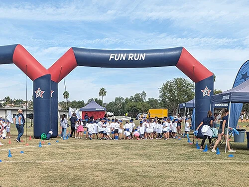 A group of children gather at the starting line under an inflatable arch labeled “Fun Run” at an outdoor event, with tents and adults nearby on a grassy field.