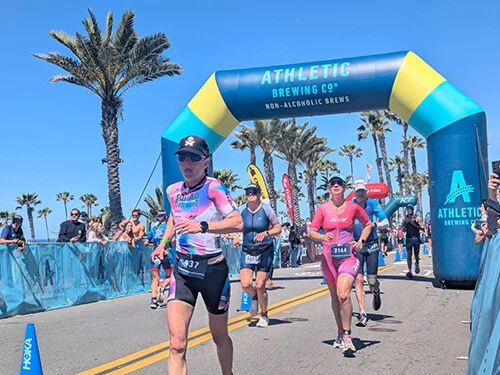 Three athletes run toward the finish line of a race under a blue and yellow archway labeled “Athletic Brewing Co. Non-Alcoholic Brews” with palm trees and spectators in the background.