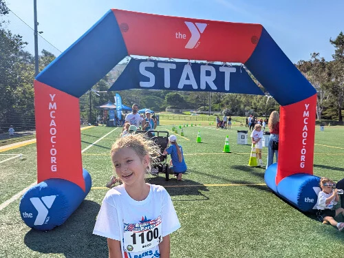 A smiling girl stands in front of a YMCA START arch on a sunny sports field, wearing a race bib. Other people and race participants are visible in the background.