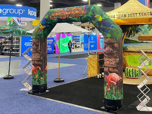 A trade show booth with a tropical-themed inflatable arch reading NOW ENTERING INSTANT PARADISE, decorated with jungle leaves, a flamingo, and tiki statues, surrounded by displays and other colorful booths.
