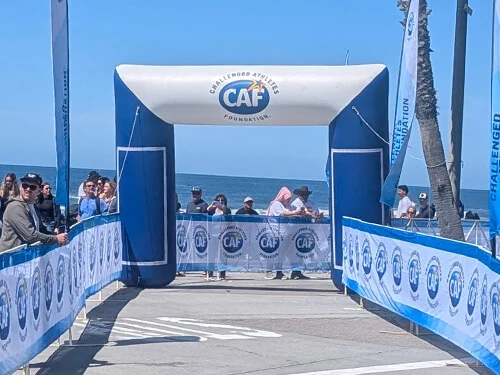 A finish line archway branded with CAF Challenged Athletes Foundation stands by the ocean, surrounded by people and blue banners on a sunny day. Spectators line both sides of the race route.