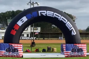 An inflatable black arch with HBM REAPER written on it stands on a grassy field. American flag-themed bases support the arch, and raceway buildings are visible in the background.
