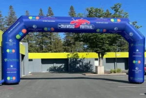 A large blue inflatable arch with Crawdad Festival and a red crawdad logo spans a parking lot entrance, surrounded by trees and closed storefronts in the background. Sponsor logos cover the arch.