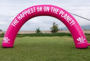 A large pink inflatable arch stands on grass, displaying the words THE HAPPIEST 5K ON THE PLANET! in white letters, promoting The Color Run event.