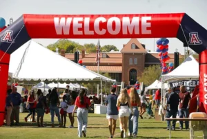 People walk under a large red “Welcome” arch at an outdoor event with tents, balloons, and a historic building in the background. Some attendees wear casual clothes, and the atmosphere is festive.