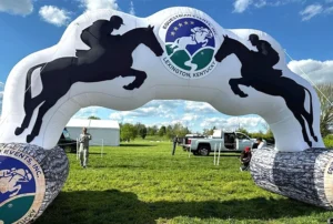 An inflatable archway features silhouettes of jumping horses and riders with the Equestrian Events Inc. Lexington, Kentucky logo. People and vehicles are visible on the grassy field under a partly cloudy sky.