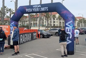 An inflatable archway with VEGA PUSH YOUR LIMITS stands at an event start or finish line. People gather nearby. Palm trees, a hotel, and parked cars are visible in the background.