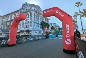 A street race finish line with two large red inflatable TriDot arches, blue cones, palm trees, and a white multi-story building in the background under a clear blue sky.