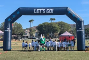 A group of children in matching shirts stands under a large inflatable arch that reads LETS GO! on a grassy field, with trees, houses, and a tent in the background.