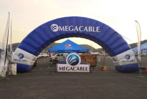 A large blue inflatable arch with the Megacable logo stands in a parking lot, with a Megacable sign below it. Cars and branded tents are visible in the background at sunset.