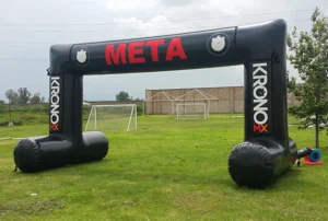 An inflatable black arch with META in red letters stands on a grassy field near soccer goals. The arch displays the KRONO MX logo and some circular icons, suggesting its used as a finish line for an event.