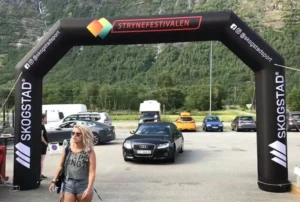 A woman in sunglasses walks under a black inflatable arch with “STRYNEFESTIVALEN” written on it. Several parked cars and a mountainous landscape are visible in the background.