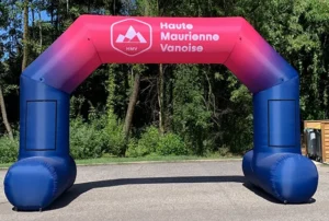 An inflatable archway in blue and pink with the logo and text Haute Maurienne Vanoise stands outdoors on a paved surface, with trees and greenery in the background.