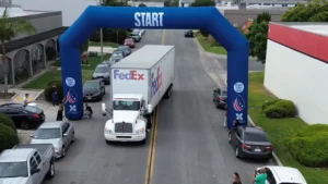 A large FedEx truck drives under a blue inflatable START archway on a street lined with parked cars and people standing nearby. The scene appears to be part of an event or race.