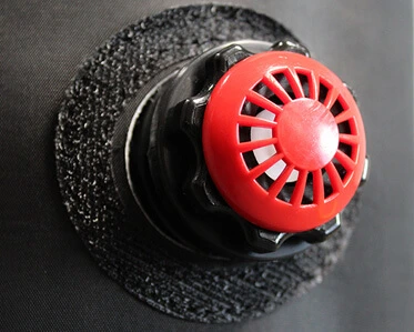 A close-up of a red air vent or valve attached to a black circular base on a dark surface. The vent has a protective grill design.