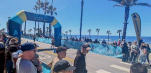 A crowd gathers near a blue inflatable archway at a beachside road event, with palm trees, a clear blue sky, and the ocean in the background. Spectators line both sides of the street, watching the activities.