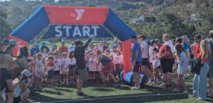 A crowd of children in running gear gather at a START line under a YMCA arch, with adults cheering and supervising on a sunny day at an outdoor event.