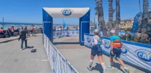 Two athletes walk hand-in-hand toward the Challenged Athletes Foundation finish gate at a sunlit seaside event, with spectators, palm trees, and ocean waves in the background.