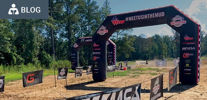 Large inflatable archways labeled #MEETUSINTHEMUD and various sponsor logos mark the starting line of an outdoor mud race event, with banners and trees in the background on a sunny day.