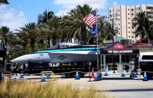A fighter jet is displayed outdoors near palm trees and buildings, with U.S. flags and a canopy labeled Top Gun Experience. Traffic cones and barriers surround the exhibit under a sunny sky.