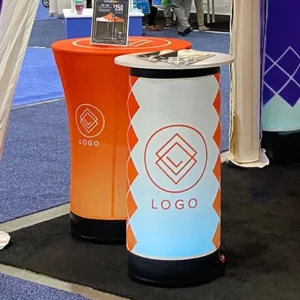 Two cylindrical display tables with LOGO text and a geometric logo, one orange and one white with orange accents, are shown at a trade show booth with carpeted blue and black flooring.