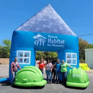 Five people stand smiling under a large blue inflatable house with Pensacola Habitat for Humanity written on it, next to two large green inflatable objects shaped like hard hats.