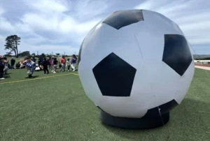 A large inflatable soccer ball sits on a green sports field, with a group of people gathered in the background under a partly cloudy sky.