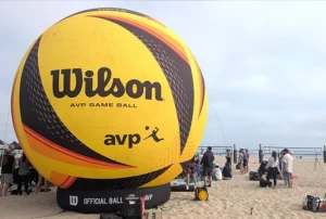 A giant inflatable Wilson AVP game ball stands on a sandy beach near a volleyball court, with groups of people gathered around and playing volleyball in the background.