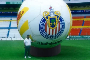 A man stands on a soccer field next to a giant soccer ball featuring the Club Deportivo Guadalajara (Chivas) logo, with stadium seats and advertisements visible in the background.