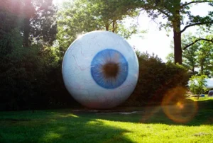 A large, realistic sculpture of a human eyeball with blue iris stands on a grassy lawn, surrounded by green trees. Sunlight shines through the trees, creating a lens flare in the image.