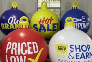 Five large, colorful holiday ornaments with promotional phrases such as PRICED LOW, Hot SALE!, COUPONS, Our BRAND, and SHOP & EARN displayed in a room with bright windows.
