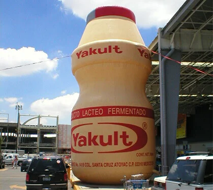 A giant inflatable Yakult bottle stands outdoors near a building and parking lot, with cars and a partly cloudy sky in the background.