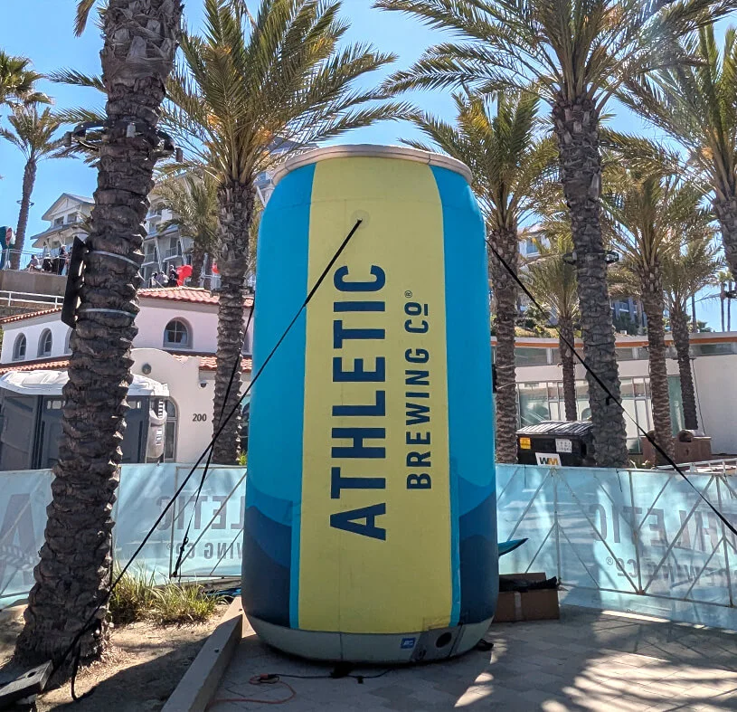 A large inflatable can shaped like a beer can with ATHLETIC BREWING CO printed on it stands among palm trees at an outdoor event on a sunny day.