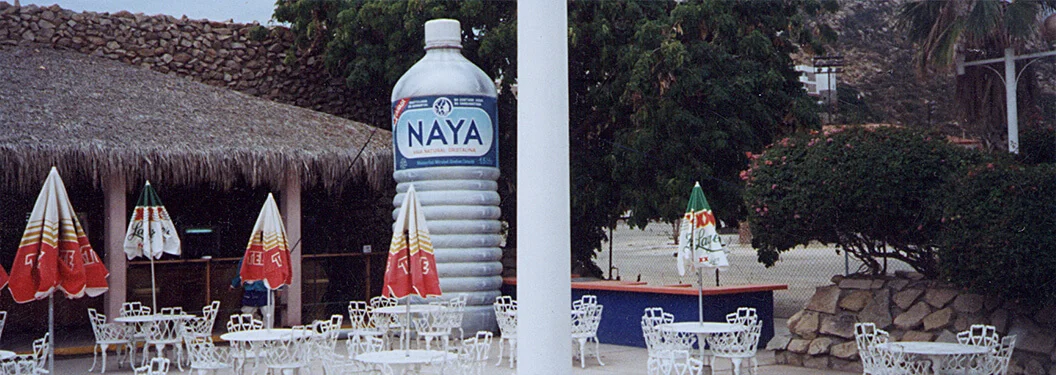 Outdoor cafe with white metal tables and chairs, colorful umbrellas, and a large, upright Naya water bottle statue near a thatched roof building, surrounded by greenery and a white pillar in the foreground.