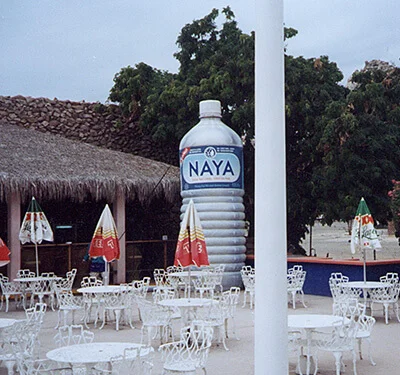 A large outdoor café area with white metal chairs and tables, colorful umbrellas, and a giant Naya water bottle replica standing upright near some trees and a thatched-roof building.