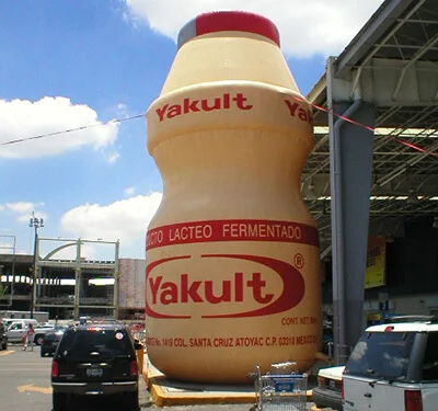 A giant replica of a Yakult probiotic drink bottle stands outdoors near a parking lot and building, surrounded by cars and people under a partly cloudy sky.